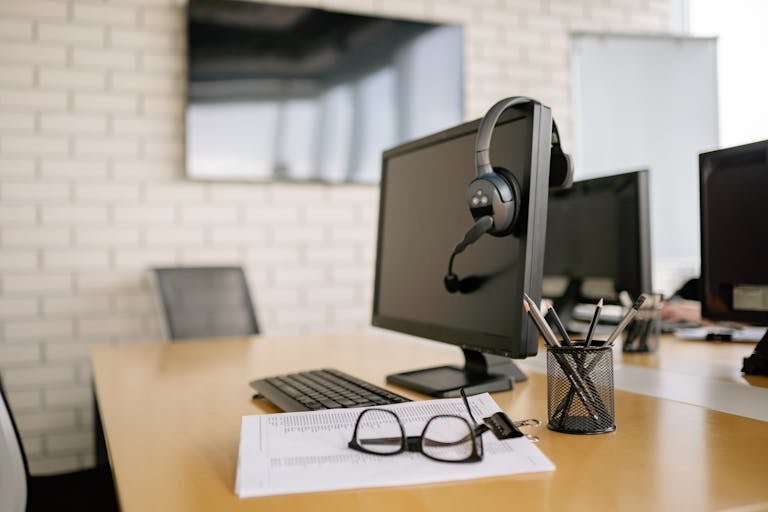 A clean modern office desk setup featuring a computer, headphones, documents, and eyeglasses.