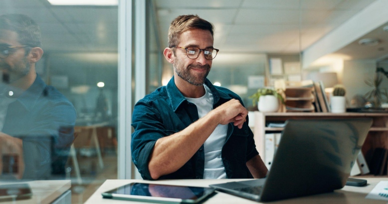 Man in blue shirt working at a desk with a laptop and plants in an office setting