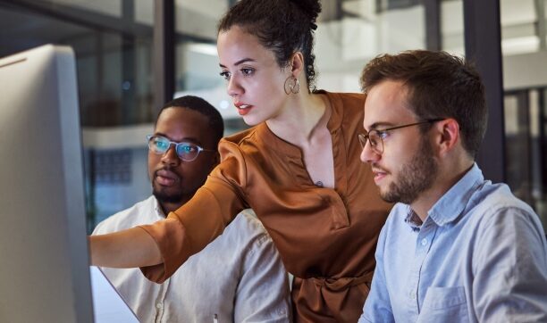 Three professionals collaborating at a computer in a modern office at night