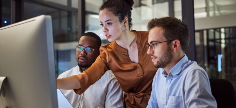 Three professionals collaborating at a computer in a modern office at night