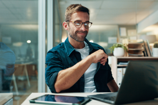 Man with glasses smiling at laptop in modern office workspace