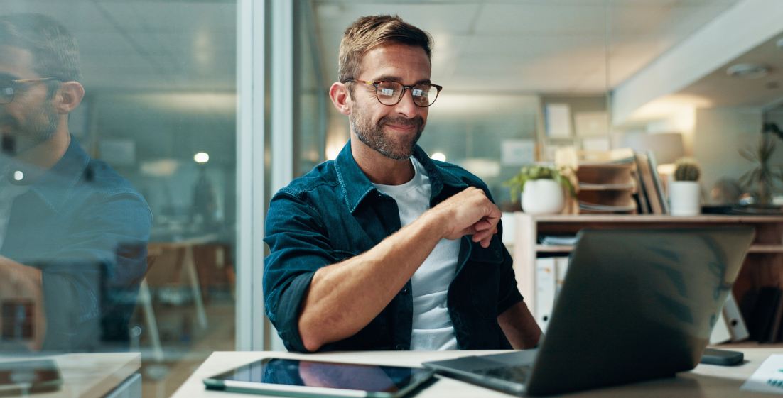 Man with glasses smiling at laptop in modern office workspace